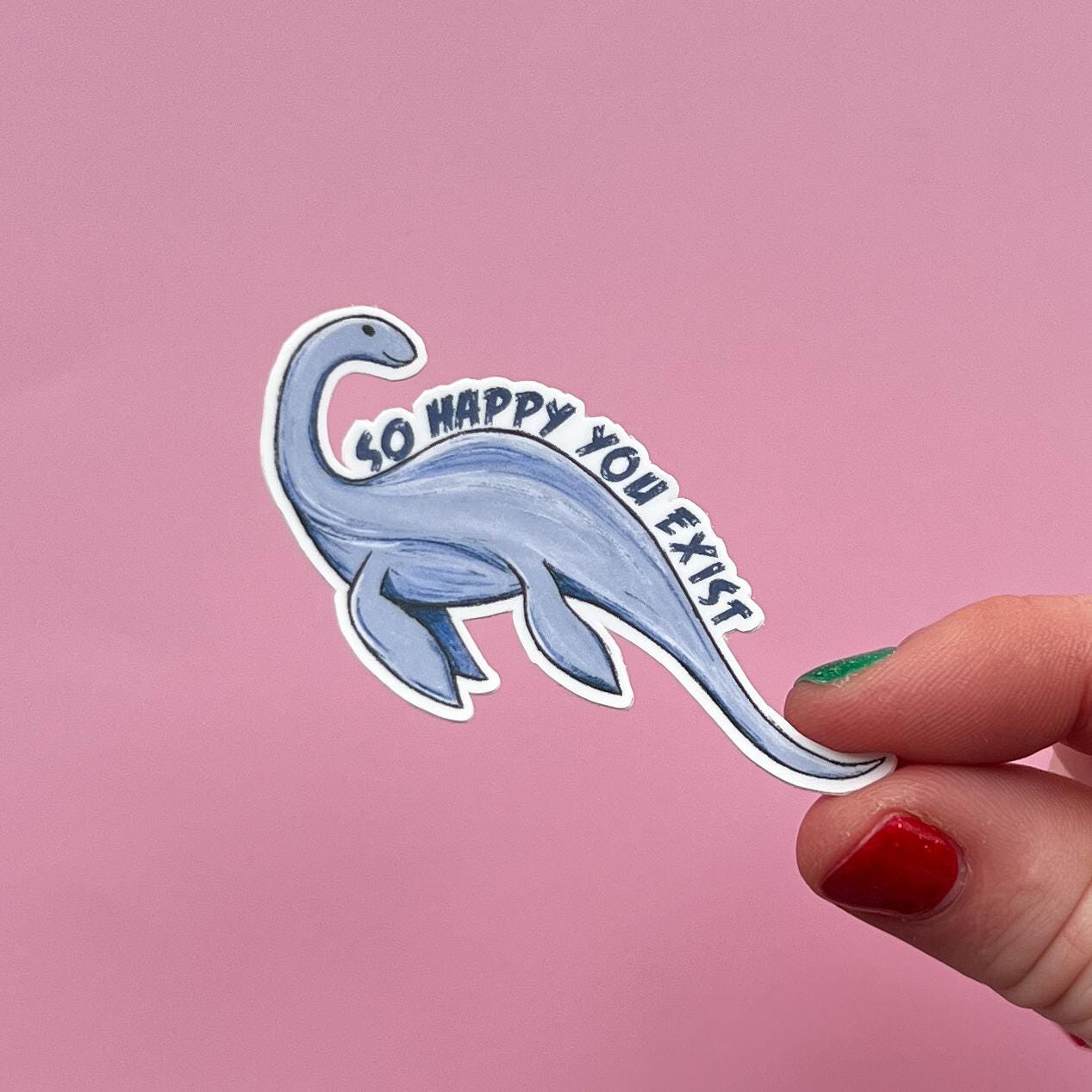 Hand holding a Loch ness monster or Champy sticker with 'So happy you exist' text on a pink background