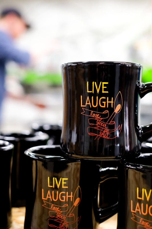 Black mug with 'Live Laugh Love Your Wife' text in orange on a reflective surface.