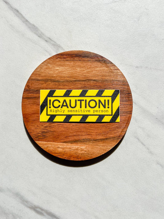 Wooden coaster with a caution sticker that says "caution highly sensitive person" on a gray surface
