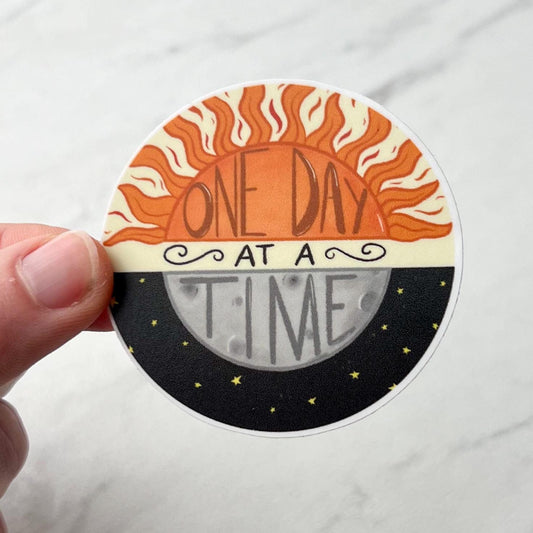 Circular sticker with sun and moon design and 'One Day at a Time' text held by a hand on a white background