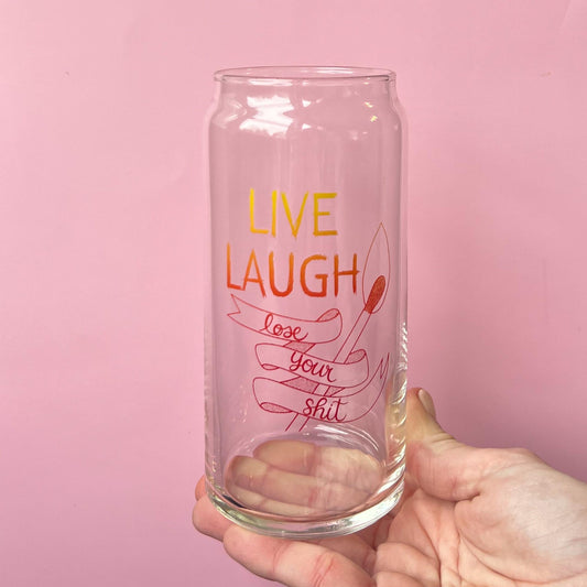 Glass tumbler with 'Live Laugh Lose Your Shit' text held against a pink background