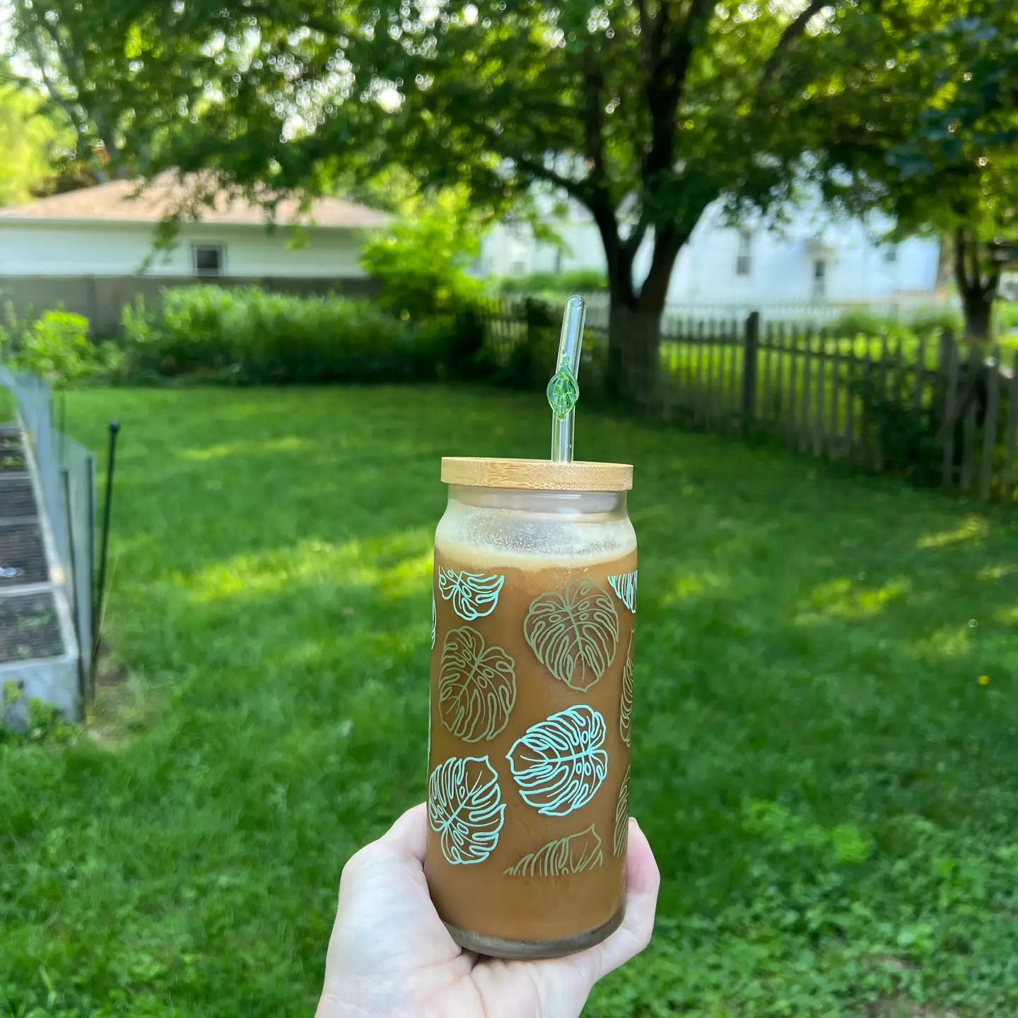 Hand holding a clear glass tumbler with monstera patterns outdoors on a grassy area.