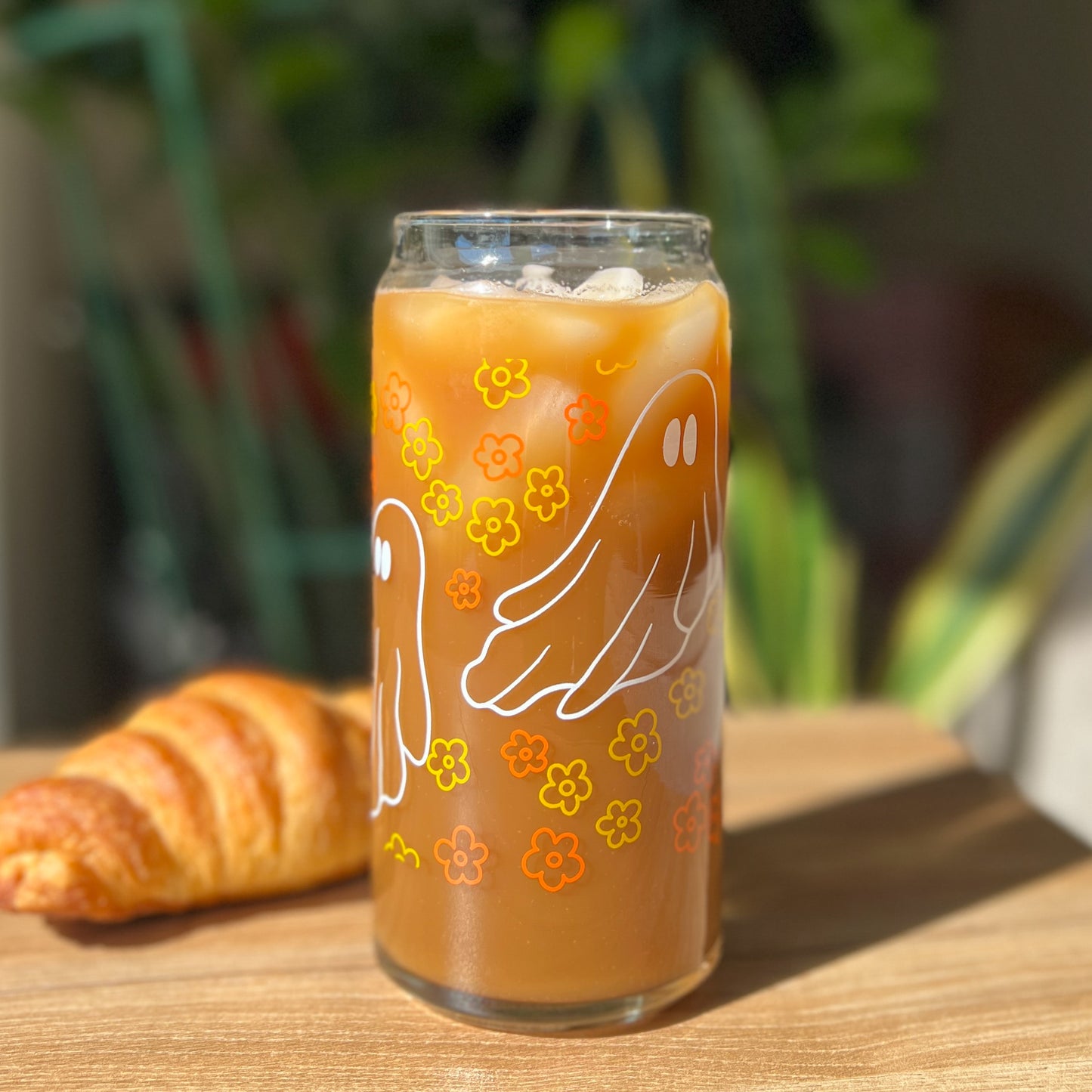 Glass of iced coffee with floral and ghost design on a wooden table with a croissant.