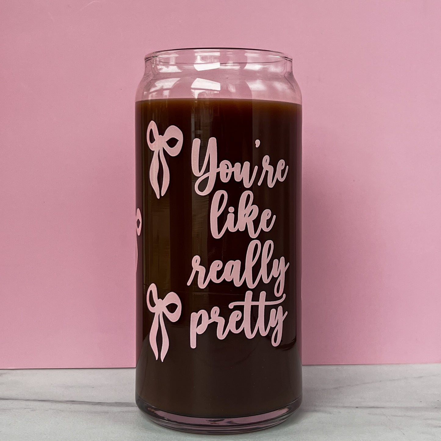 Glass cup with 'You're like really pretty' text and pink ribbons on a pink background