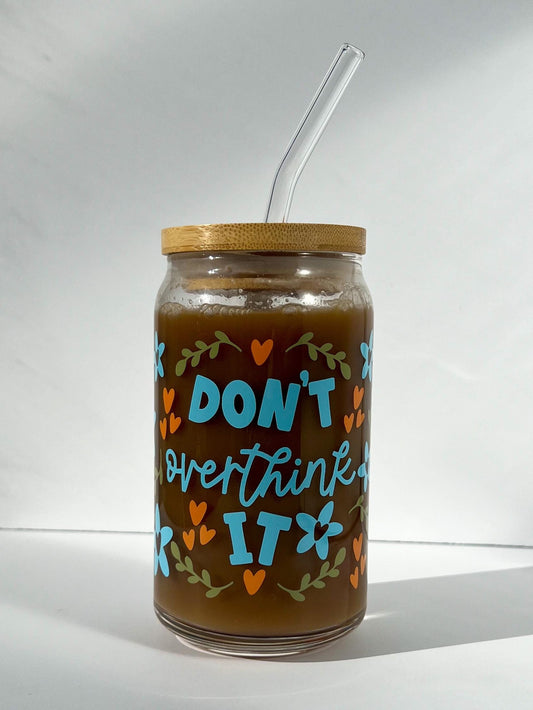 Glass jar that says "don't overthink it" with wooden lid and straw, containing a beverage, on a white background