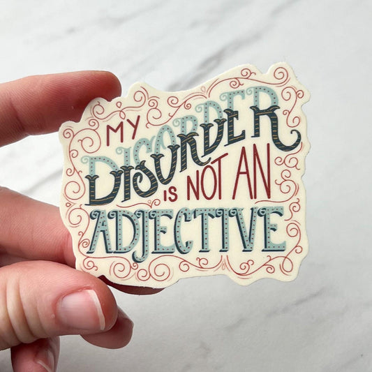 Hand holding a decorative sticker with text 'My Disorder is Not an Adjective' against a white background