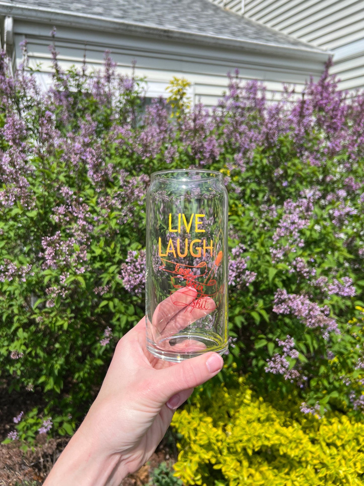 Clear tumbler with 'Live Laugh Lose your shit' text held in front of a garden with purple flowers.