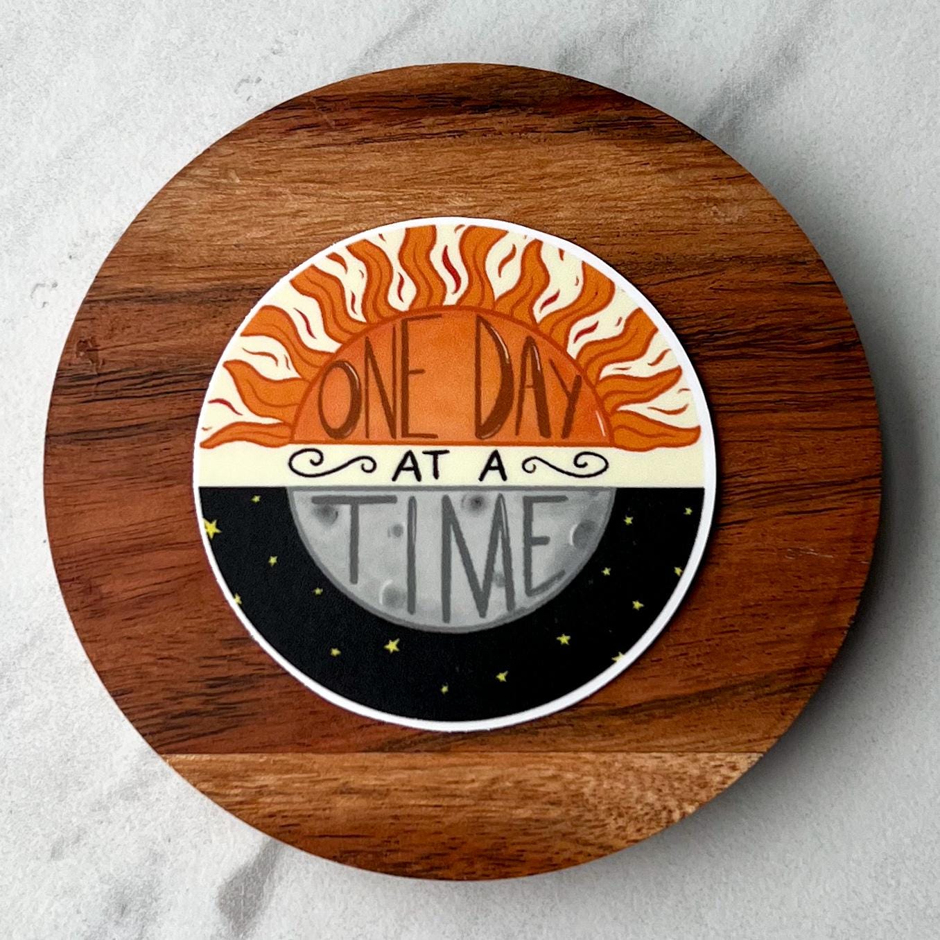 Circular sticker with sun and moon design and 'One Day at a Time' text 