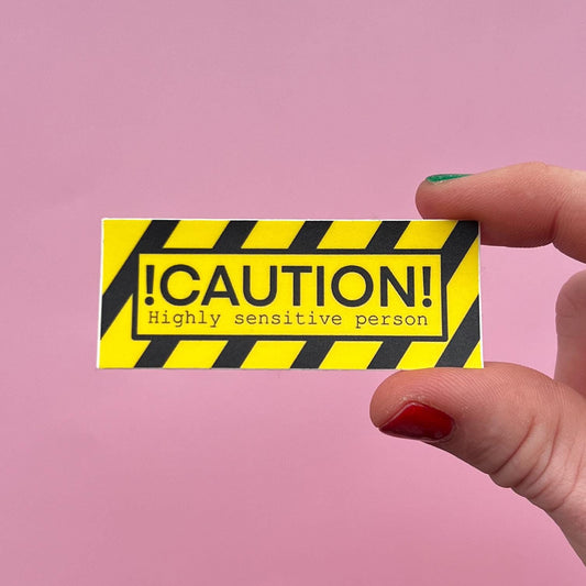 Hand holding a yellow and black caution label with 'Caution: Highly sensitive person' text on a pink background