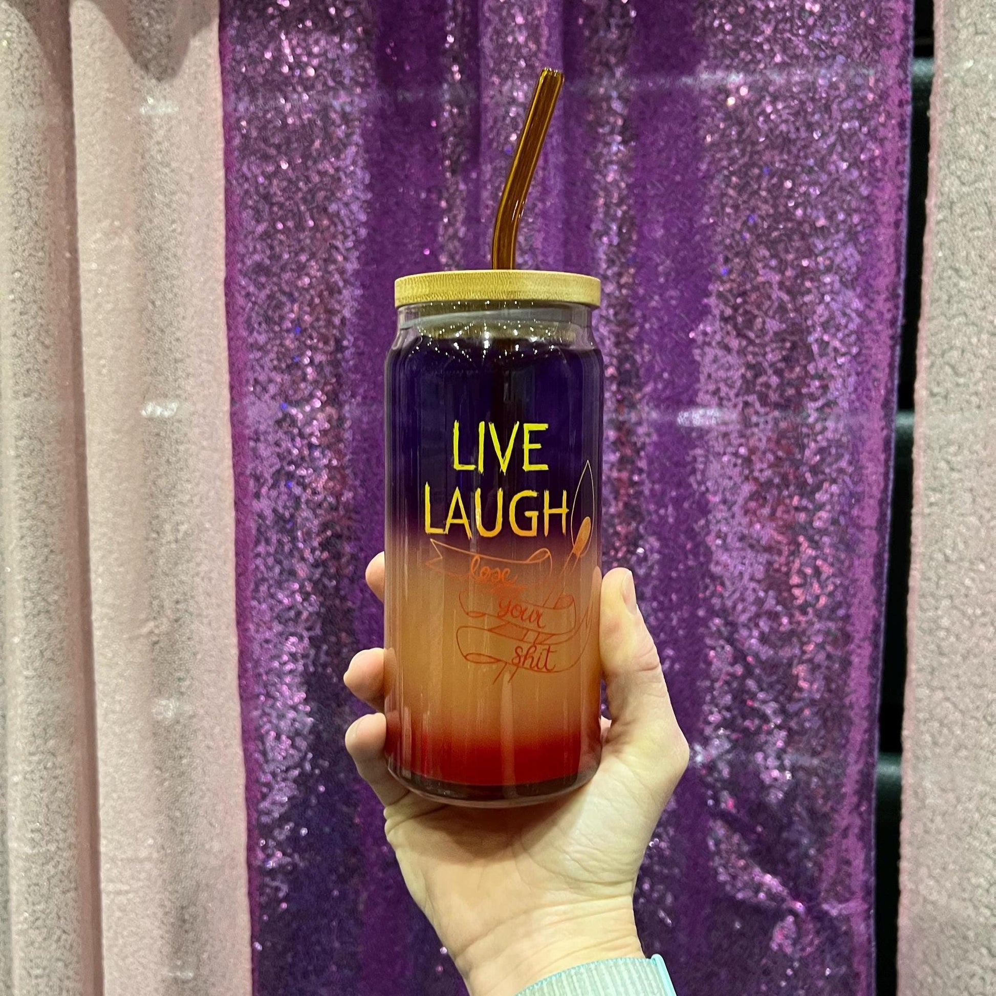 Hand holding a colorful tumbler with 'Live Laugh' text against a purple glittery background