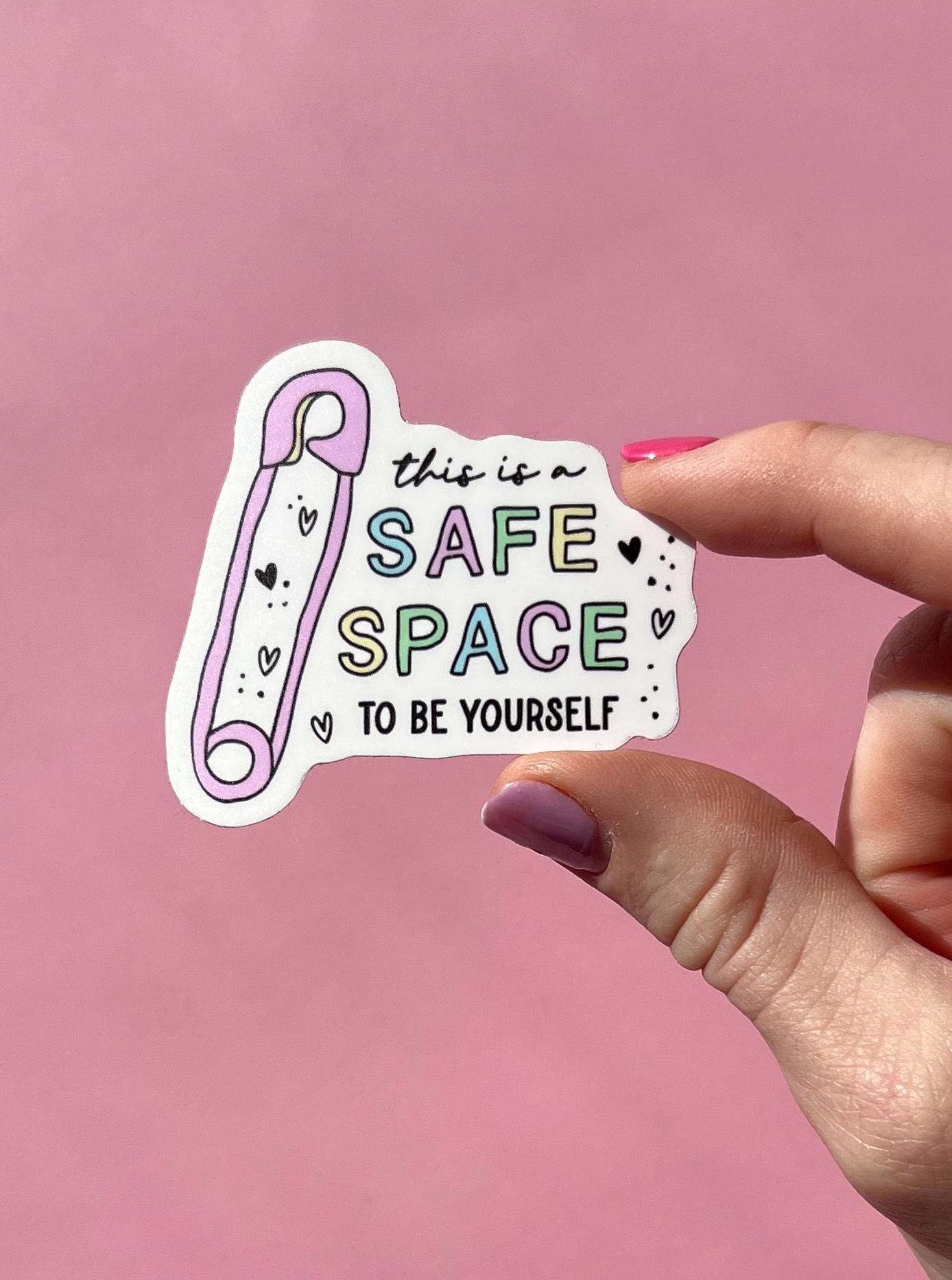 Hand holding a sticker with 'This is a safe space to be yourself' text on a pink background