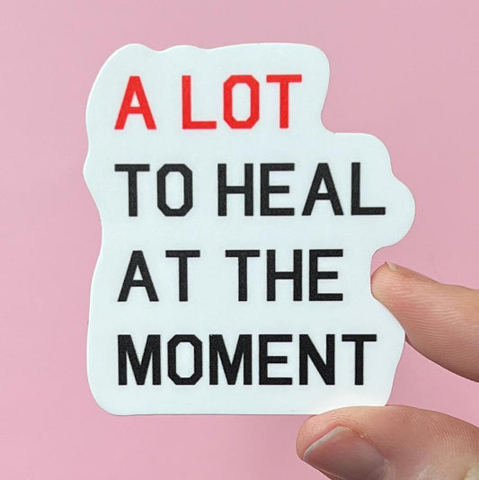 Sticker with text 'A LOT TO HEAL AT THE MOMENT' held against a pink background