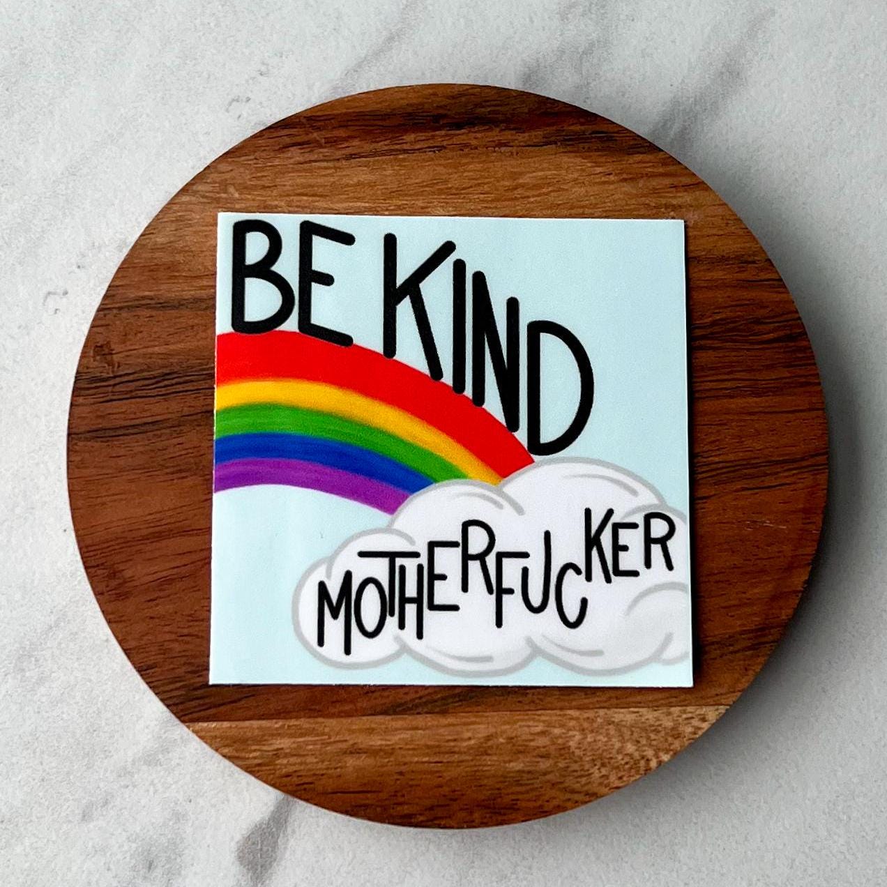 Round wooden with sticker that says 'be kind motherfucker' with a colorful rainbow on a light background
