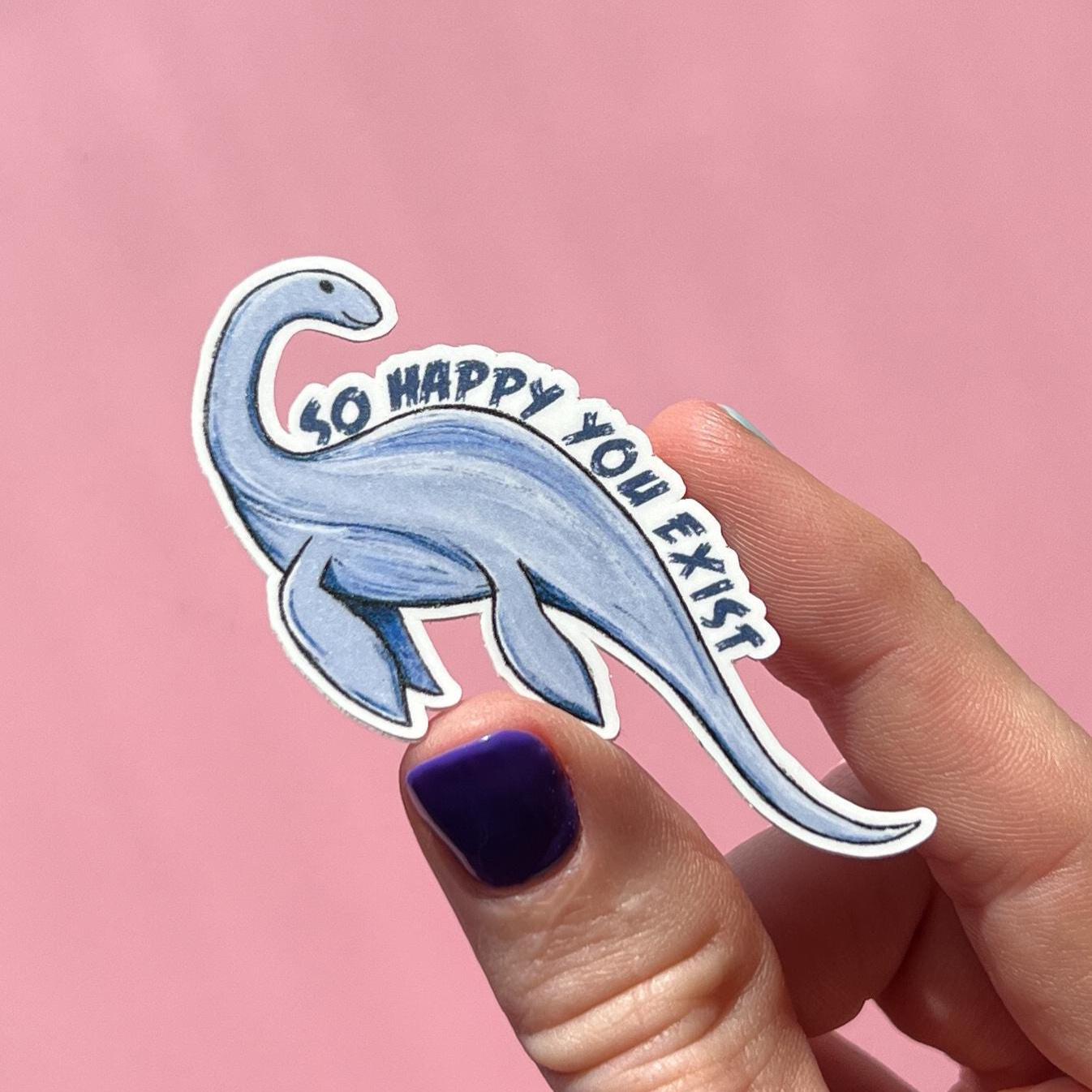 Hand holding a  Loch ness monster or Champy sticker with 'So happy you exist' text on a pink background