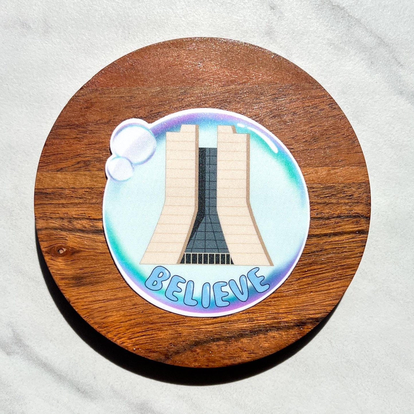 Sticker with fermilab and 'BELIEVE' text in a bubble on a wooden circle, placed on a white fabric background.