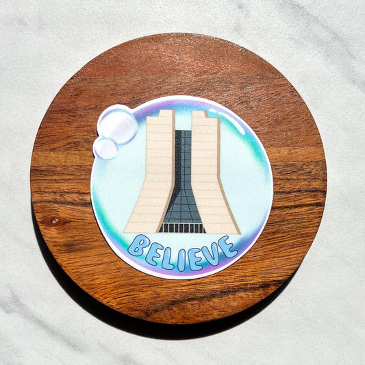 Sticker with fermilab and 'BELIEVE' text in a bubble on a wooden circle, placed on a white fabric background.