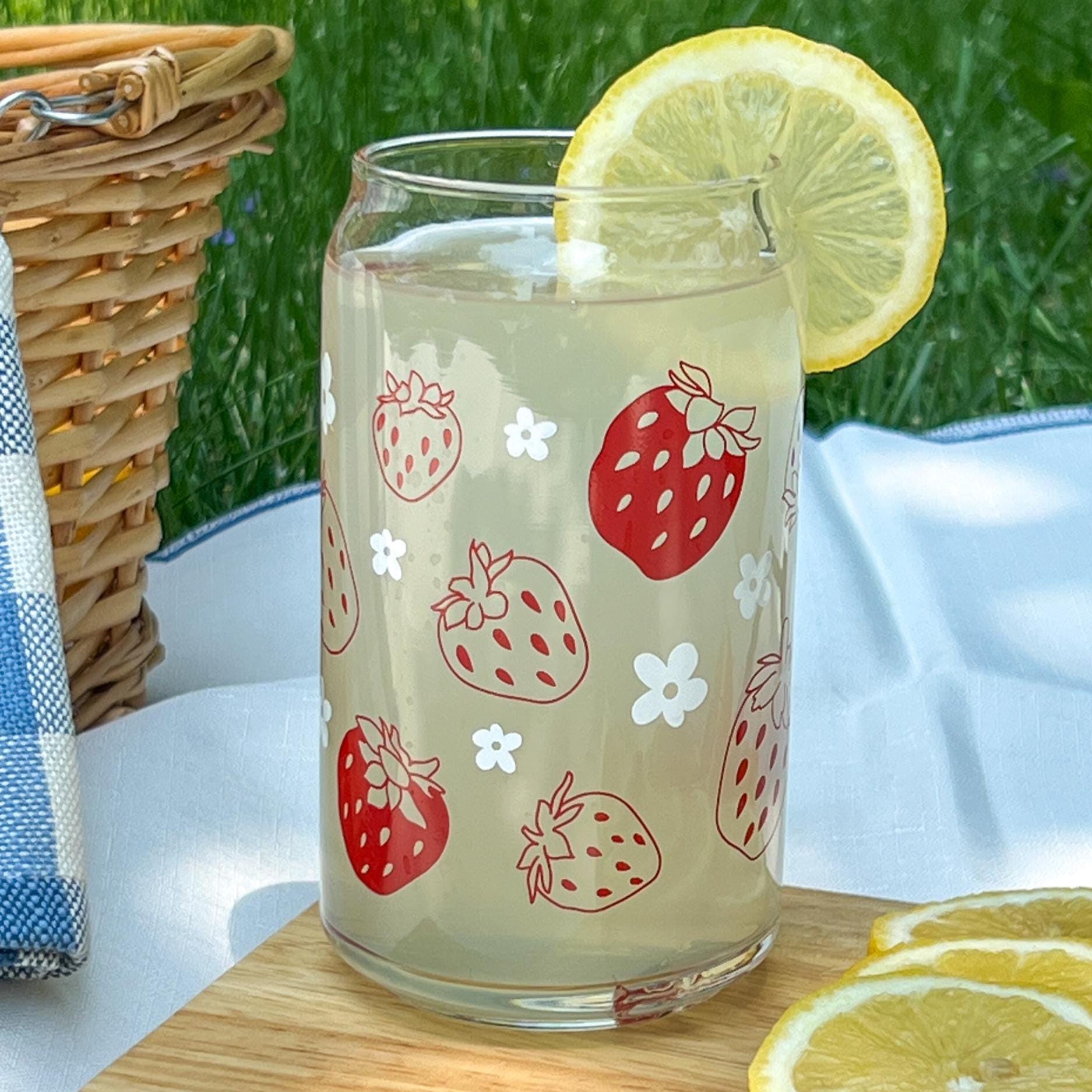 Glass with strawberry and white daisy design filled with a lemony drink, garnished with a lemon slice, on a wooden surface with grass in the background.