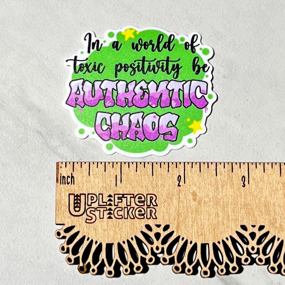 Sticker with motivational text 'In a world of toxic positivity be Authentic Chaos' on a gray background with a ruler for scale.