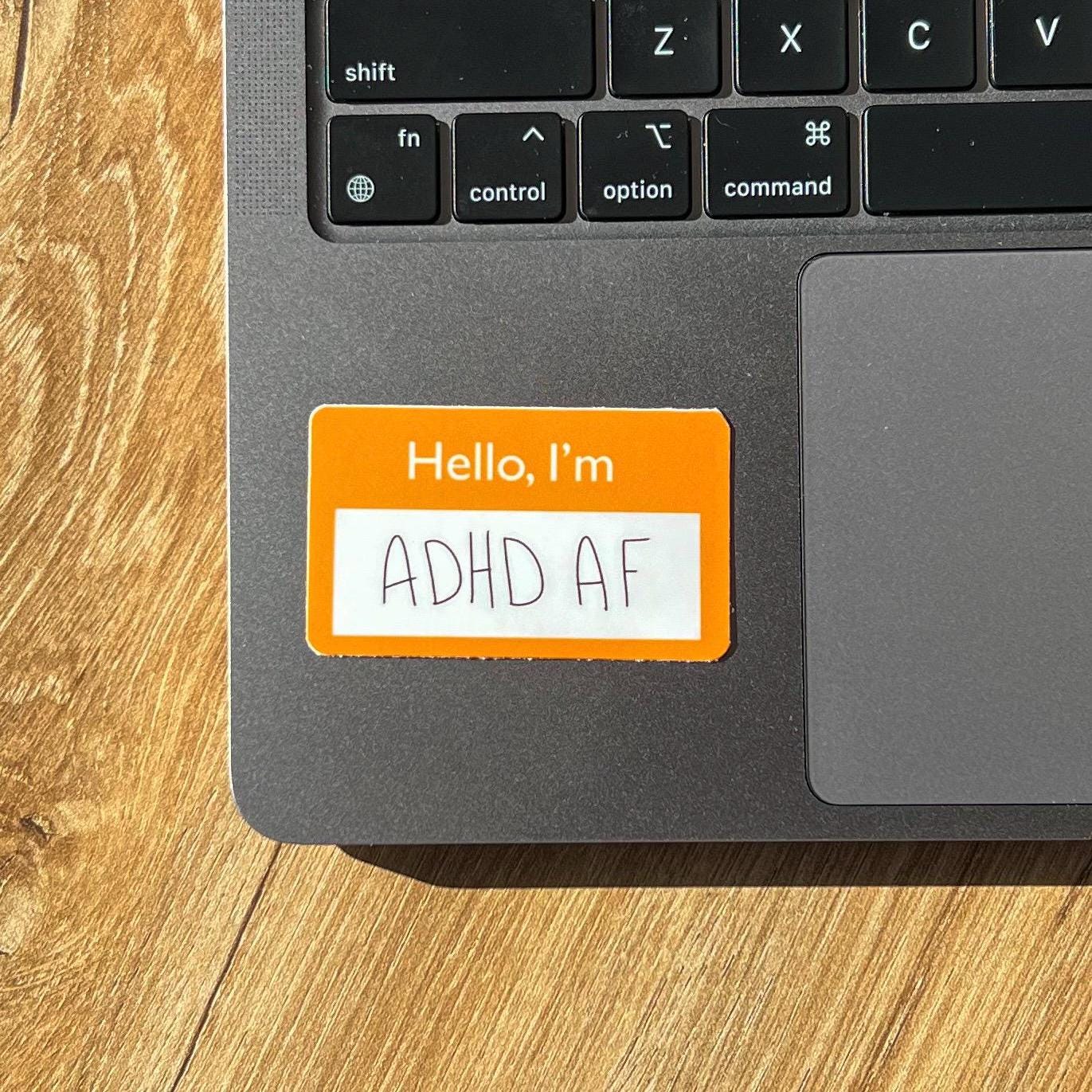 Laptop with a sticker next to the keyboard area reading 'Hello, I'm ADHD AF' on a wooden surface.