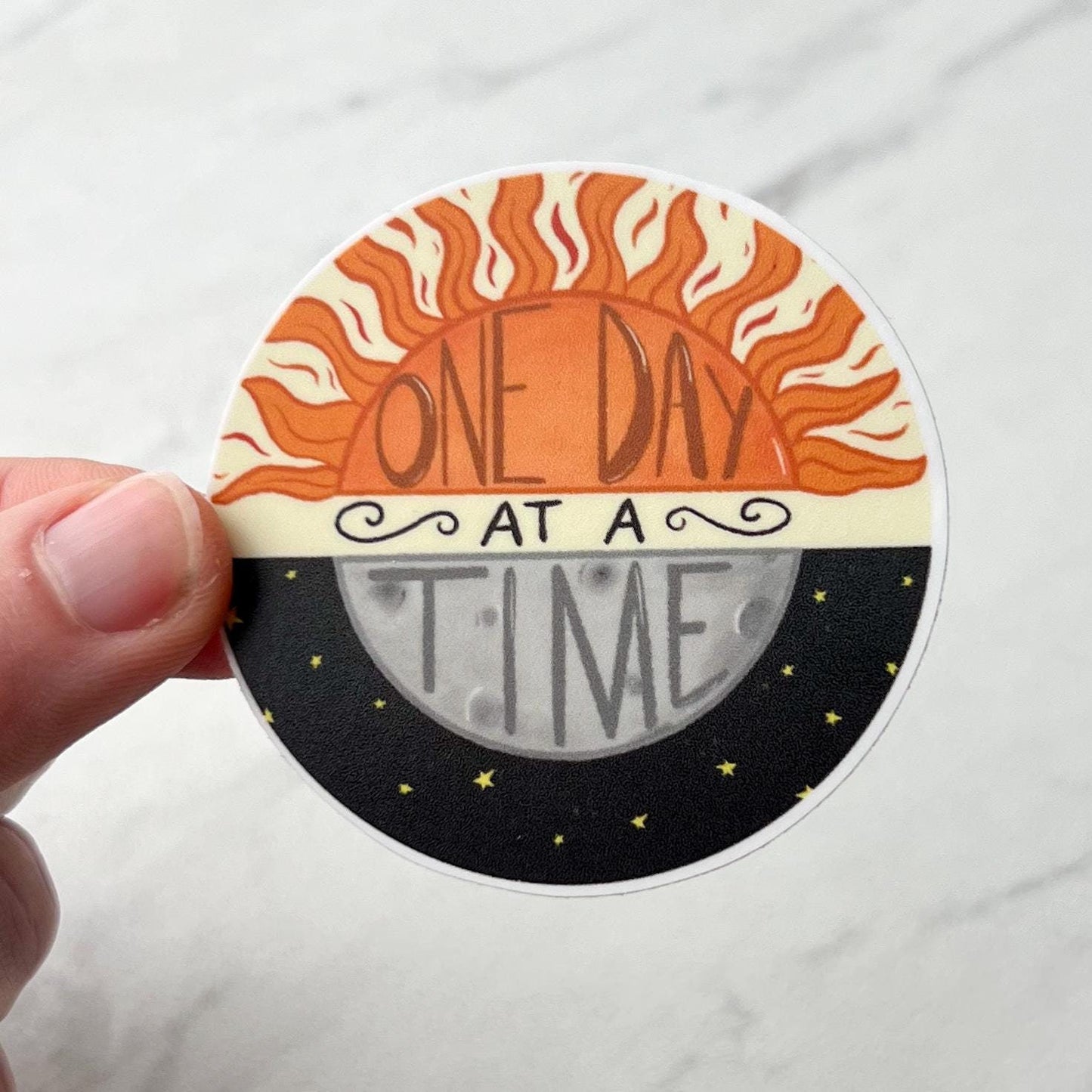 Circular sticker with sun and moon design and 'One Day at a Time' text held by a hand on a white background