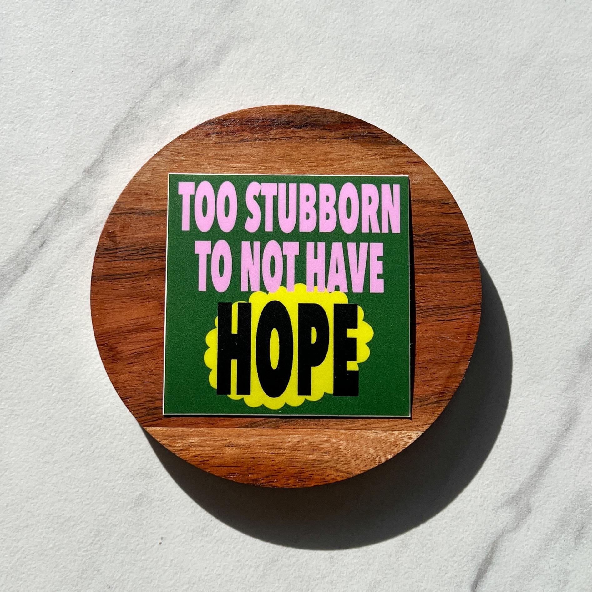 Round wooden coaster with a sticker that reads "too stubborn to not have hope" on a light gray background