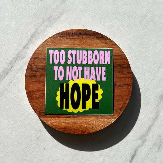 Round wooden coaster with a sticker that reads "too stubborn to not have hope" on a light gray background