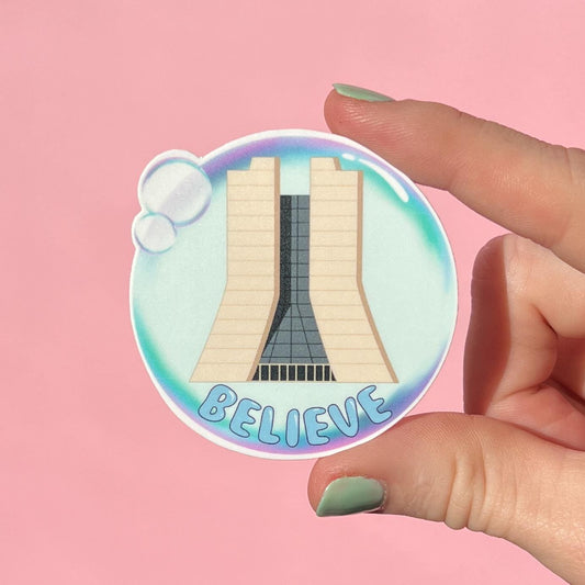 Sticker with Fermilab and 'BELIEVE' text in a bubble design held by a hand on a pink background
