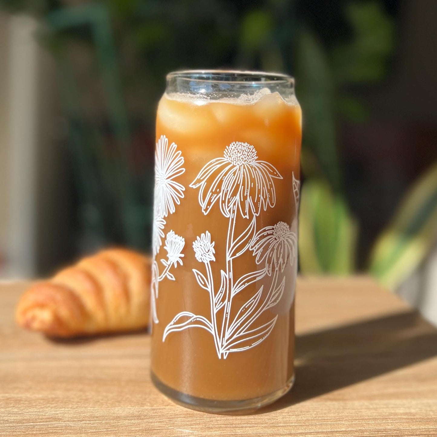 Glass of iced coffee with floral design on a wooden table with a croissant and blurred plants in the background.