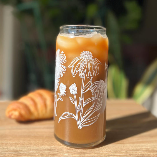Glass of iced coffee with floral design on a wooden table with a croissant and blurred plants in the background.