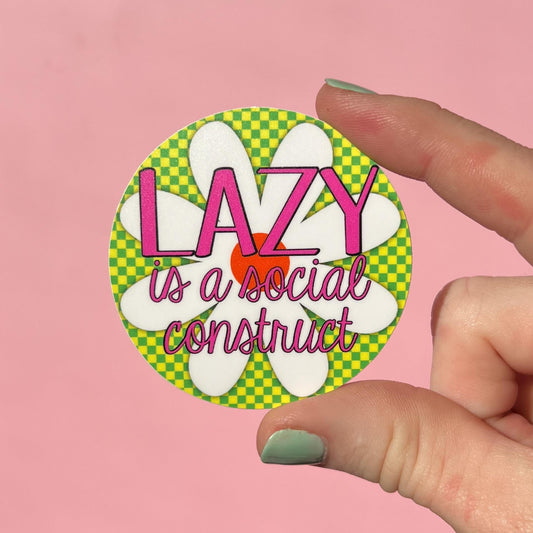 Circular sticker with 'LAZY is a social construct' text held by a hand on a pink background