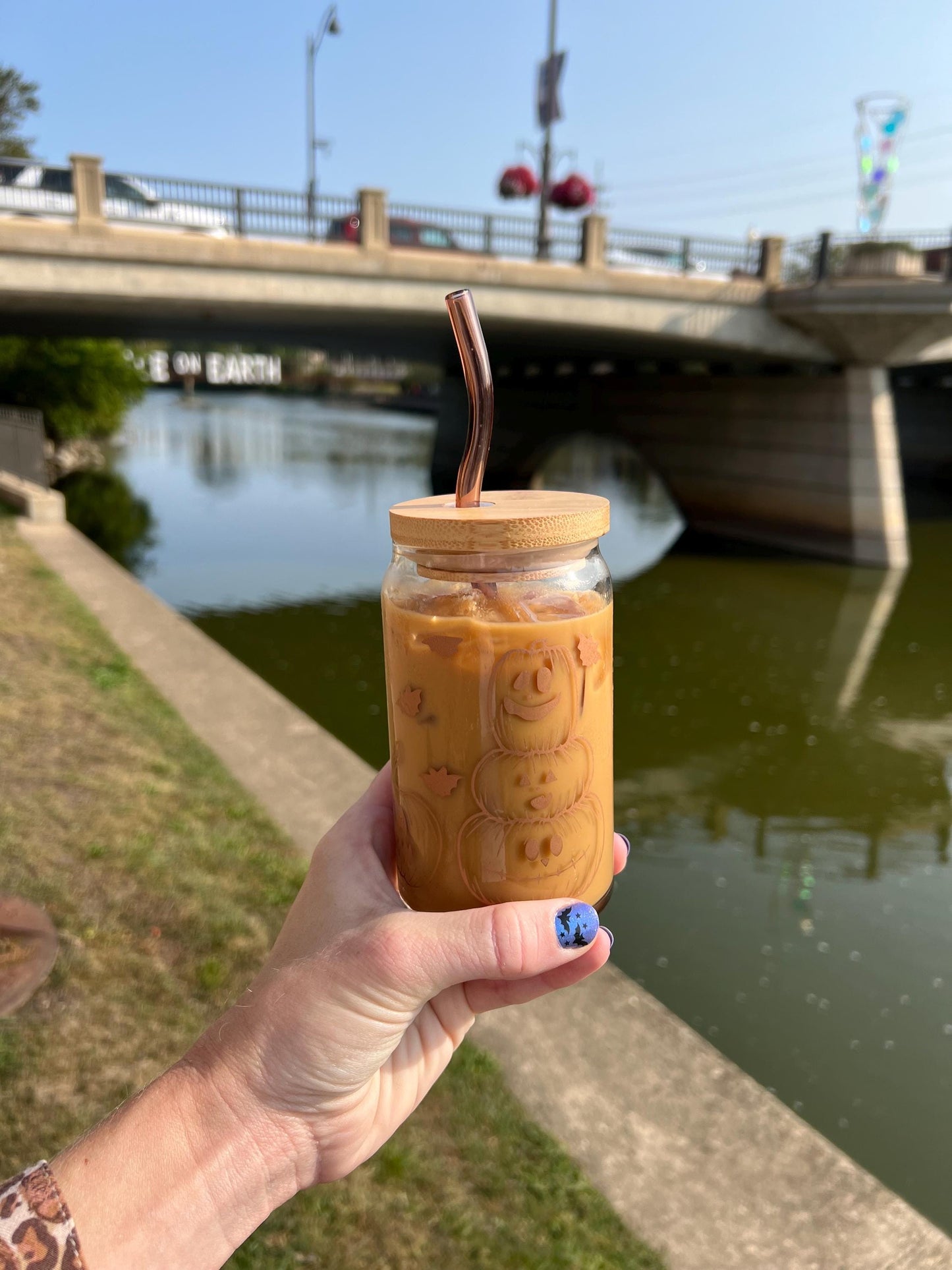 Hand holding a glass with jack o lantern and halloween themed design with a straw by a river with a bridge in the background