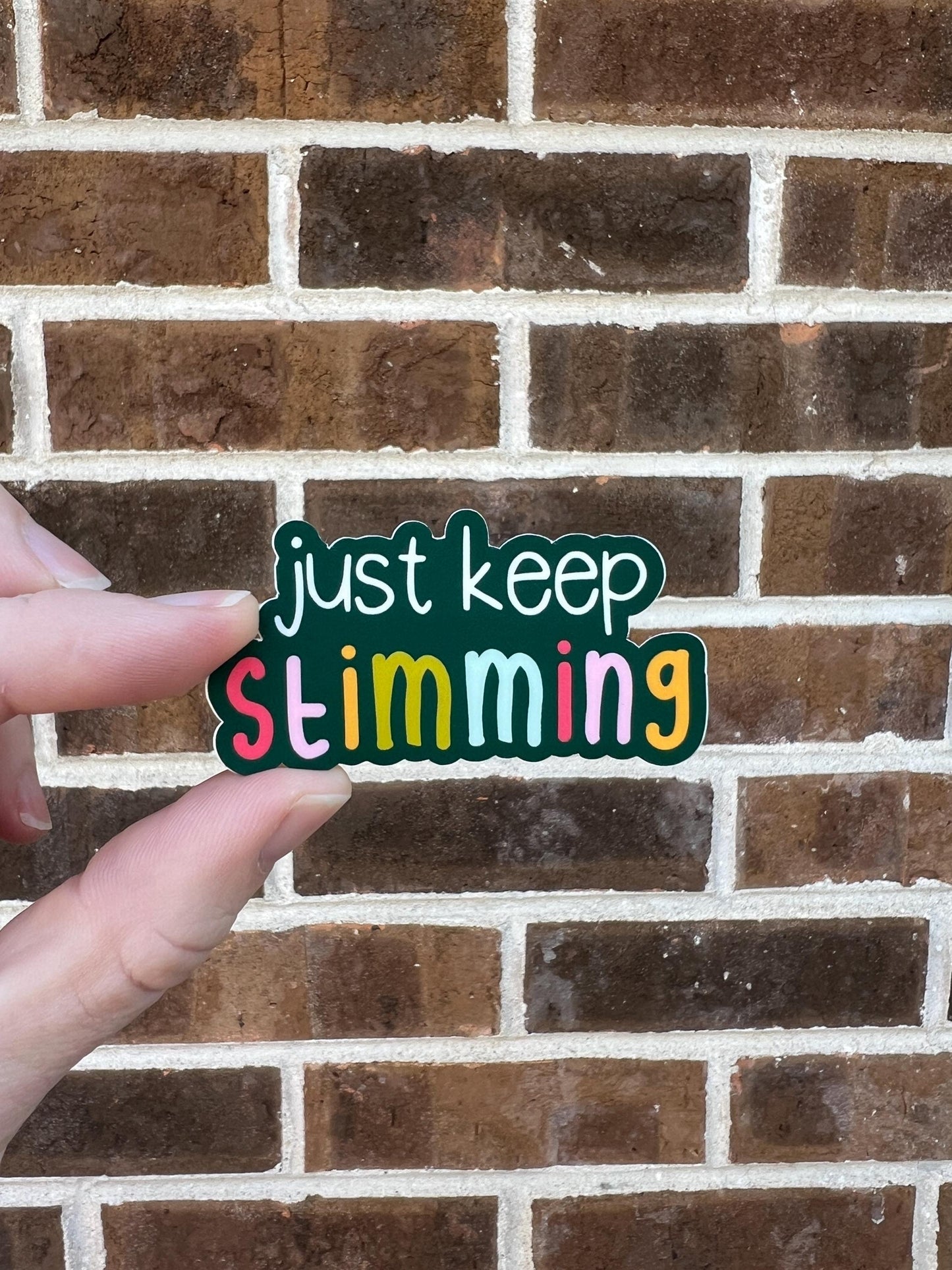 Hand holding a colorful sticker with 'just keep Stimming' text against a brick wall.
