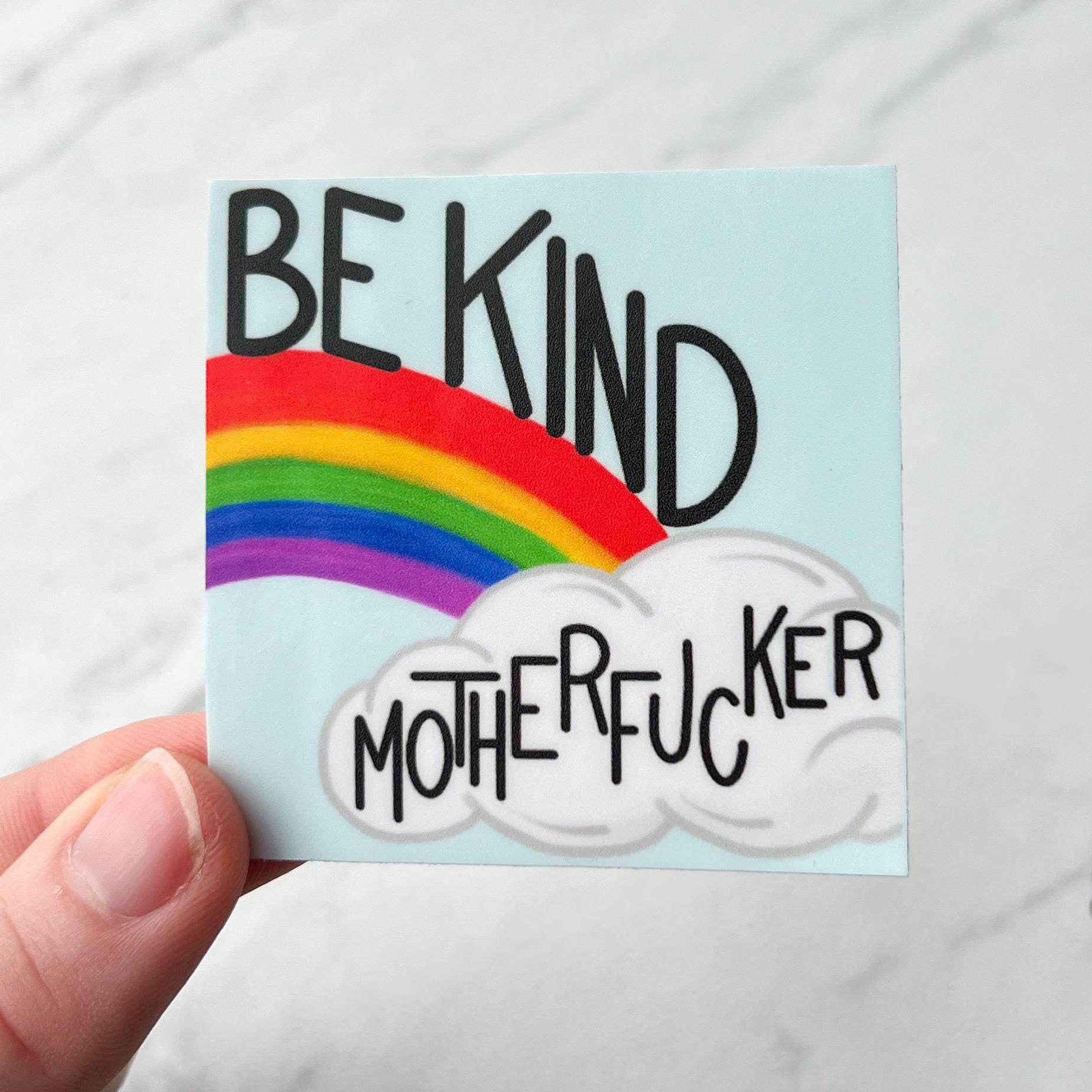 Sticker with rainbow and cloud design with text 'BE KIND MOTHERFUCKER' held by a hand.