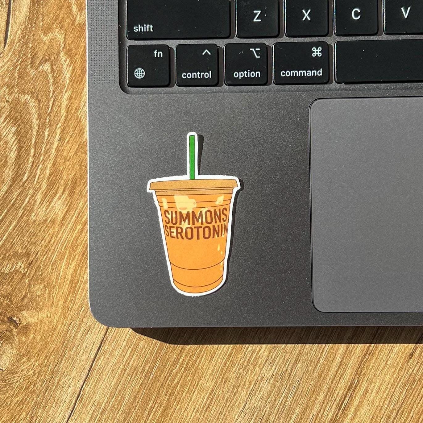 Laptop with a drink cup sticker labeled 'Summons Serotonin' on a wooden surface