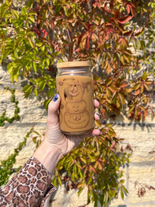 Hand holding a clear glass cup with jack o lanterns and halloween themed decorations with a lid against a leafy background
