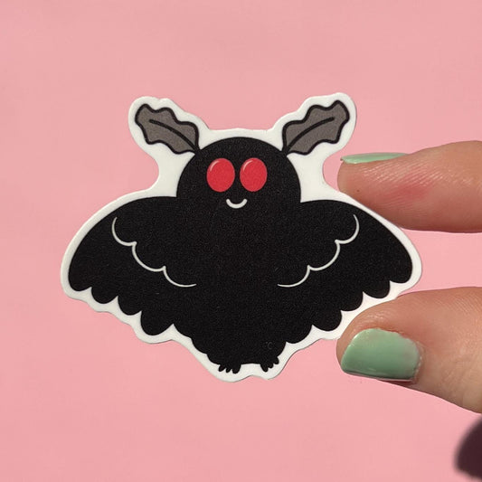 Hand holding a cute black mothman sticker with red eyes on a pink background
