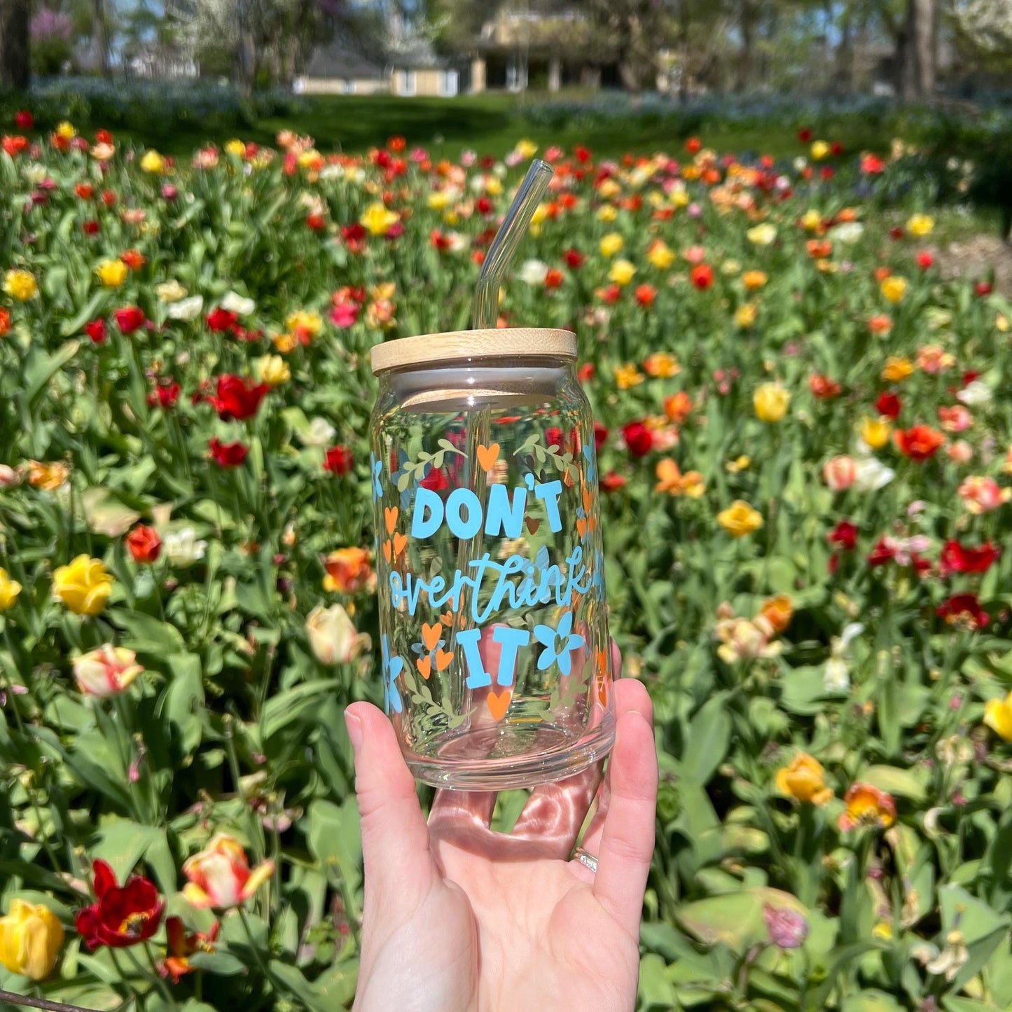 Hand holding a glass tumbler that says "don't overthink it" with a straw in front of a field of colorful flowers