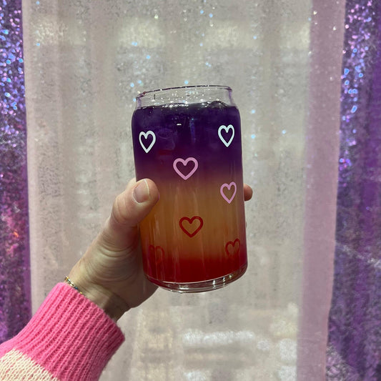 Hand holding a gradient drink in a glass with white, pink, and red heart designs against a glittery purple and white background.