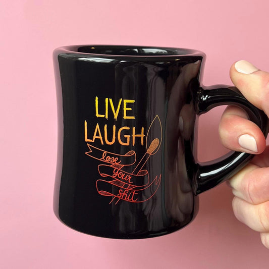 Black mug with 'Live Laugh Love Your Shit' text held by a hand against a pink background