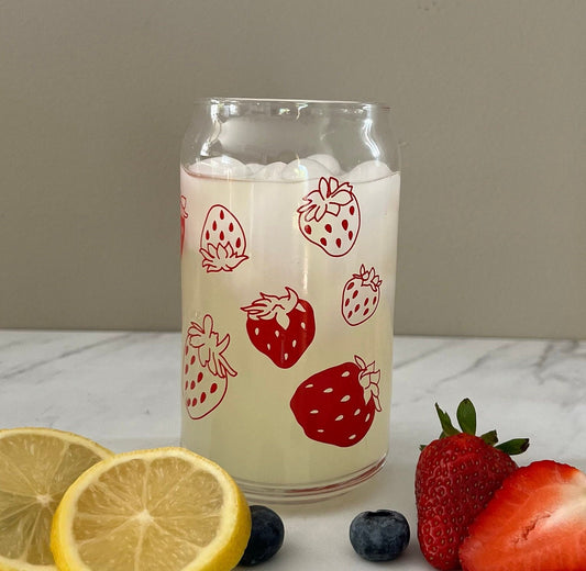 Glass with strawberry design filled with a creamy drink, surrounded by lemons, blueberries, and strawberries on a marble surface.