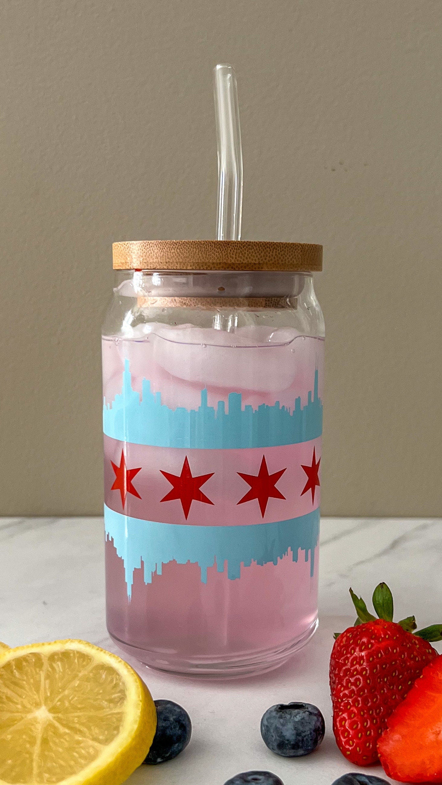 Glass jar with the Chicago flag with a blue skyline and red star design, wooden lid, straw, lemon, blueberries, and strawberries on a white surface.