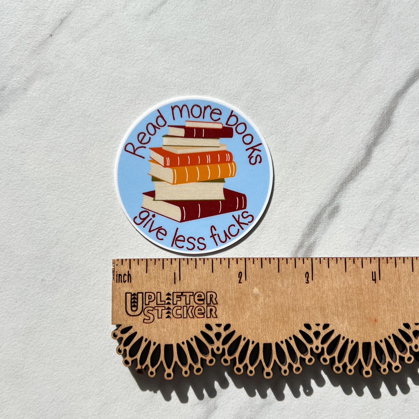 Sticker with books and text 'Read more books, give less fcks' on a gray fabric background with a ruler for scale.