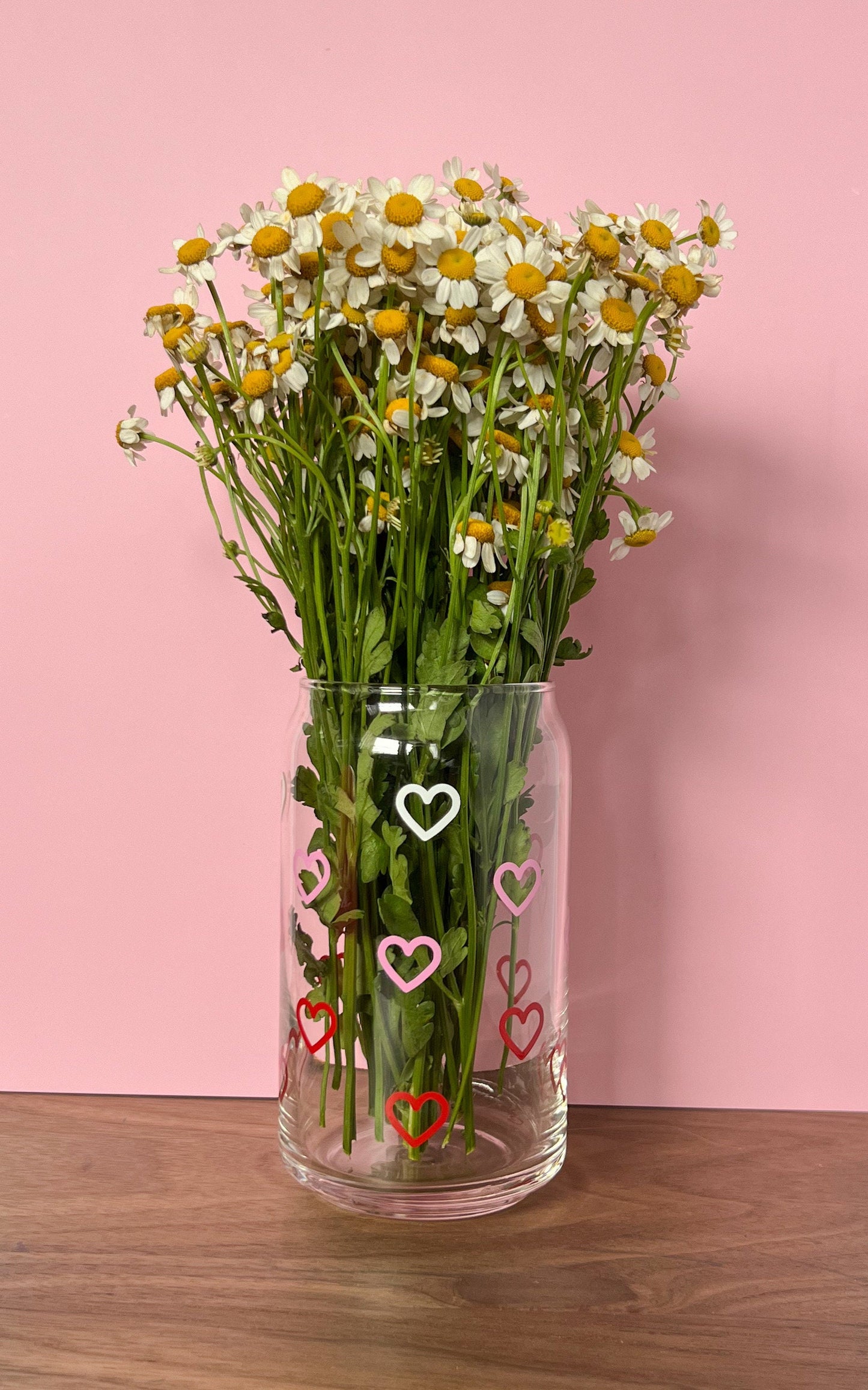 Clear vase with white, pink, and red heart designs containing a bouquet of flowers on a pink background