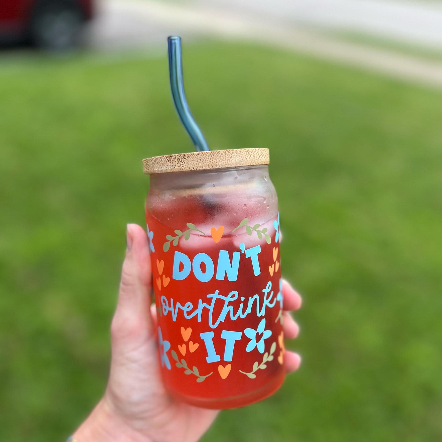 Hand holding a red drink in a glass jar with decorative text stating "don't overthink it" and straw, against a grassy background.