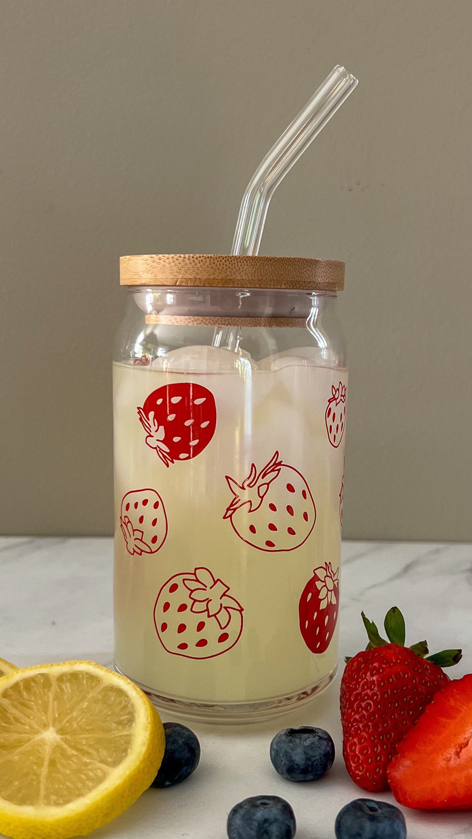 Glass jar with straw, wooden lid, and strawberry design on a white surface with fruits.