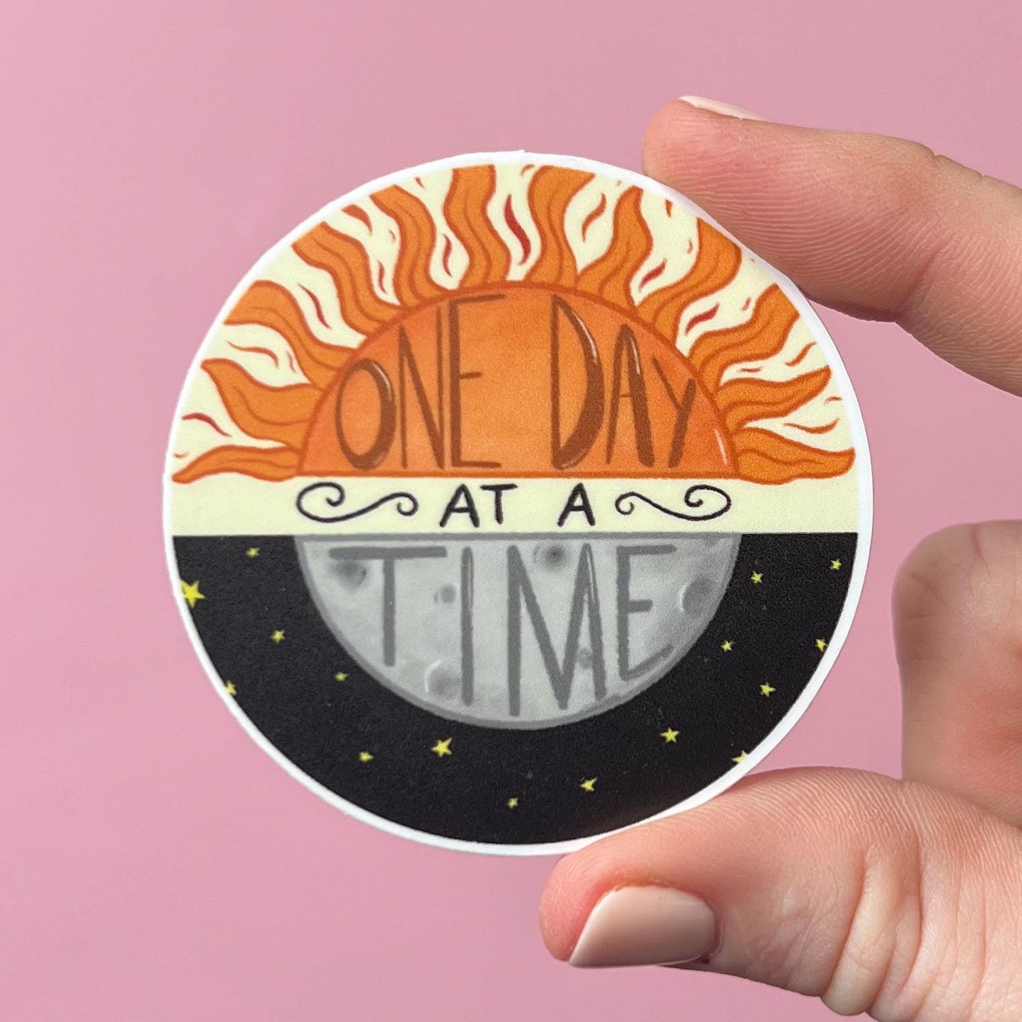 Circular sticker with sun and moon design and 'One Day at a Time' text held by a hand against a pink background