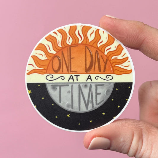 Circular sticker with sun and moon design and 'One Day at a Time' text held by a hand against a pink background