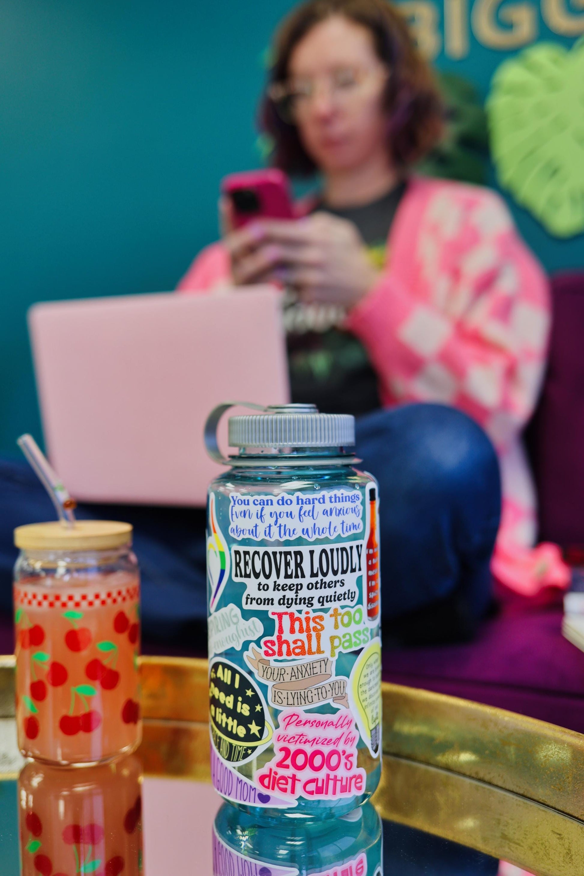 Bottle labeled 'RECOVER LOUDLY' with colorful design, person using phone in background