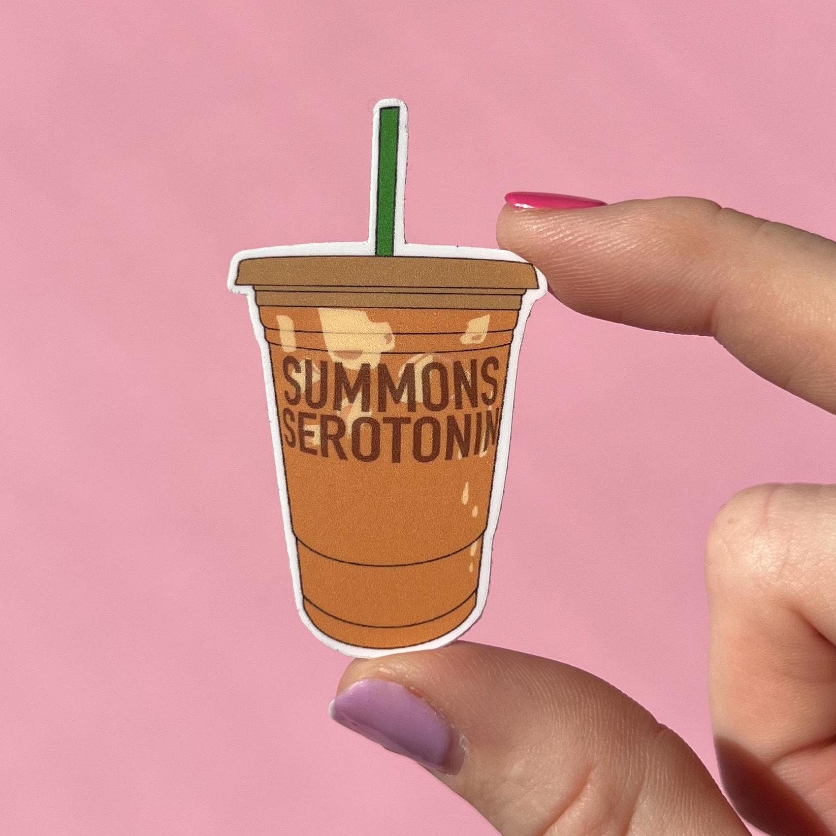 Sticker of a coffee cup with 'Summons Serotonin' text held by a hand against a pink background
