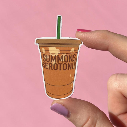 Sticker of a coffee cup with 'Summons Serotonin' text held by a hand against a pink background